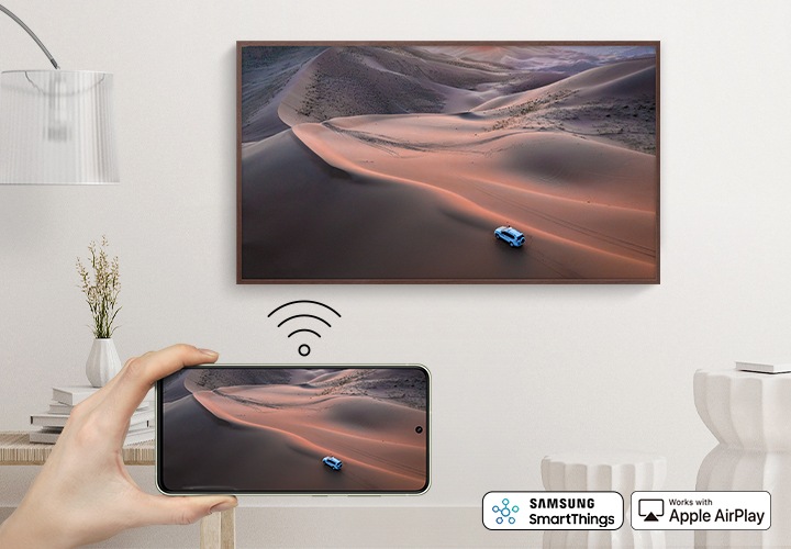 벽걸이 TV 가 설치되어 있고 앞 쪽으로는 사용자의 손과 스마트폰 화면이 보입니다.