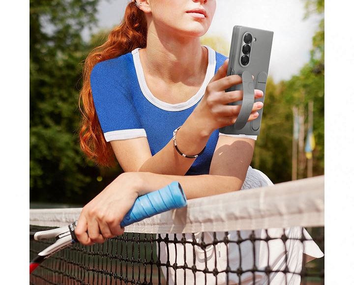 여성이 스마트폰을 편안하게 들고 테니스 코트 네트에 기대어 있습니다. 그레이 색상의 실리콘 케이스를 장착하고 있으며, 여성은 손가락에 케이스 그립을 끼워 편안하게 잡고 있습니다. 