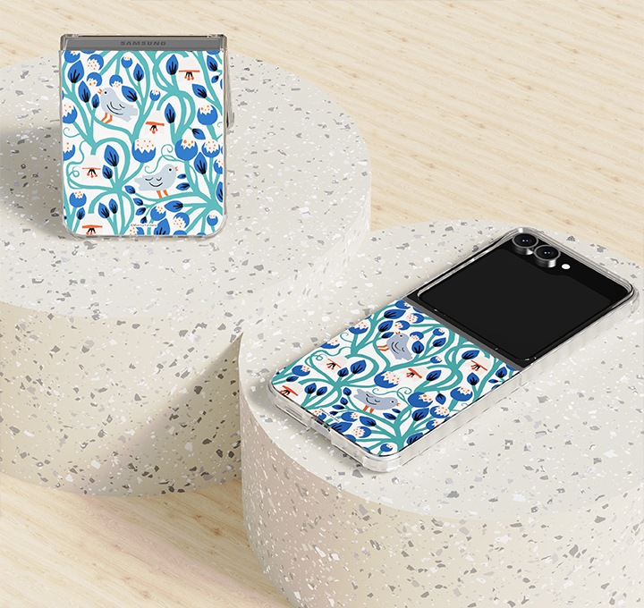 연한 나무로 된 책상을 배경으로 동그란 대리석 단상 2개가 왼쪽 상단부분에서 오른쪽 하단 부분으로 나열되어 있고 각각 왼쪽에 있는 단상 위에는 갤럭시 Z 플립6 아티스트 모니카 포스버그 버라이어티 케이스를 장착 후 90도 정도 접어 세워 놓은 정면 이미지가 보이고 있고 오른쪽 부분에 있는 단상에는 갤럭시 Z 플립6 아티스트 모니카 포스버그 버라이어티 케이스를 장착 후 펼쳐 누워있는 정면 이미지가 보이고 있습니다.