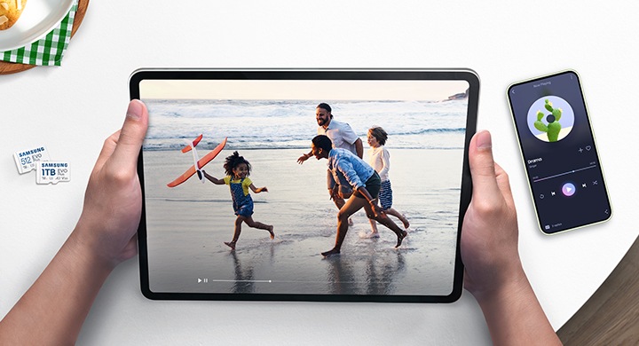 흰색의 책상을 배경으로 이미지 왼쪽 부분에는 메모리카드 정면 모습이 보이고 있고 이미지 중간 부분에는 영상을 재생하고 있는 태블릿을 양손으로 잡고 있는 이미지, 이미지 오른쪽 부분에는 스마트폰에 음악을 재생하고 있는 이미지가 보이고 있습니다.