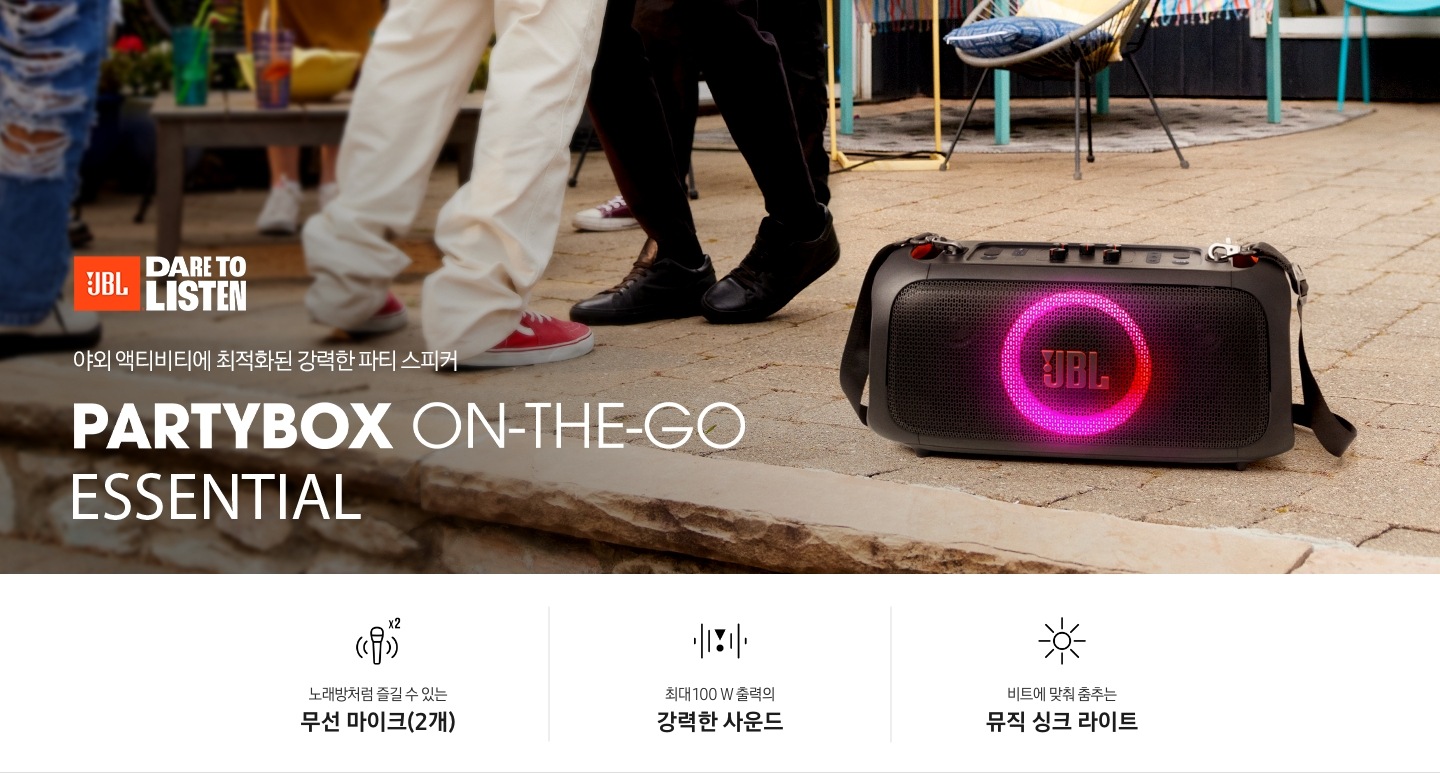 야외에 제품이 놓여 있습니다. 야외 액티비티에 최적화된 강력한 파티 스피커 PARTYBOX ON-THE-GO ESSNTIAL KV입니다. 하단에는 노래방처럼 즐길 수 있는 무선 마이크(2개), 최대 100 W 출력의 강력한 사운드, 비트에 맞춰 춤추는 뮤직 싱크 라이트 문구와 아이콘이 있습니다.