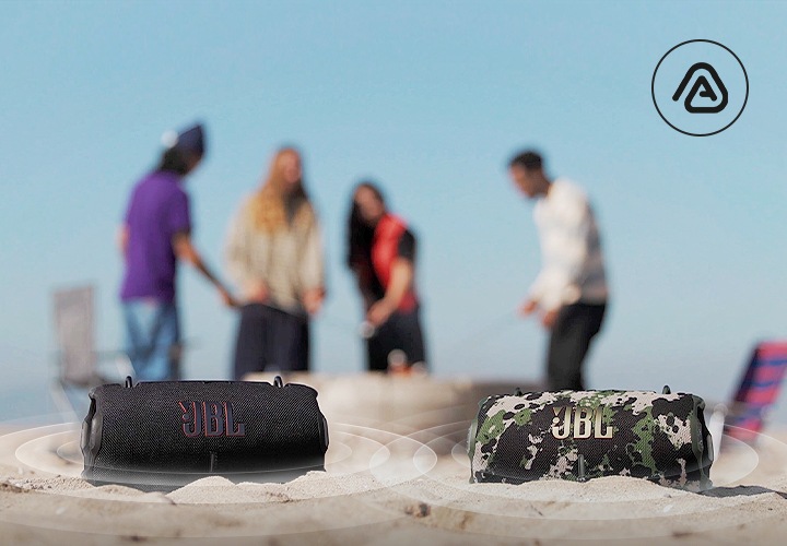 모래사장 위에 제품 2개가 놓여 있으며, 제품에서 음향효과가 나오고 있습니다. 제품 뒤로는 사람들이 서 있습니다.