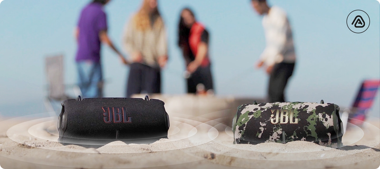 모래사장 위에 제품 2개가 놓여 있으며, 제품에서 음향효과가 나오고 있습니다. 제품 뒤로는 사람들이 서 있습니다.
