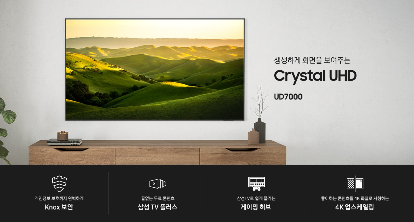 벽에 TV가 설치되어 있으며, 화면에는 푸른 산이 보입니다. 생생하게 화면을 보여주는 Crystal UHD UD7000 KV입니다. 하단에는 개인정보 보호까지 완벽하게 Knox 보안, 끝없는 무료 콘텐츠 삼성 TV 플러스, 삼성TV로 쉽게 즐기는 게이밍 허브, 좋아하는 콘텐츠를 4K 화질로 시청하는 4K 업스케일링 문구와 아이콘이 있습니다.