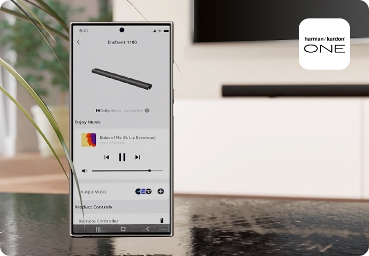 스마트폰에 Harman Kardon One 앱 화면이 보입니다. 스마트폰 뒤로는 TV와 사운드바가 흐릿하게 보입니다.