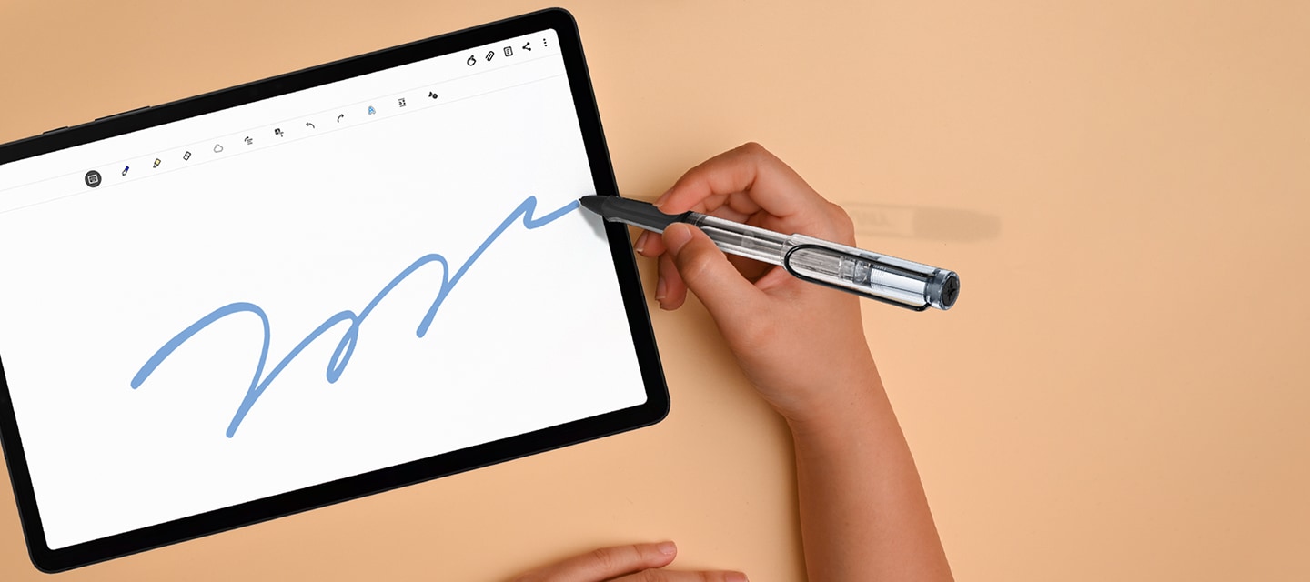 연한 주황색 책상을 배경으로 라미 사파리 S펜을 사용하여 탭에 그림을 그리고 있는 사람의 손을 위에서 내려본 이미지입니다.