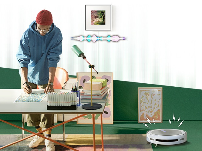 남성이 책상 위에서 작업을 하고 있습니다. 남성 뒤 벽에 뮤직 프레임이 설치되어 있으며, 하단에는 그래픽 효과가 보입니다. 책상 오른쪽에는 무선 로봇 청소기가 청소 중에 있습니다.