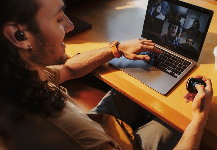 귀에 이어폰을 착용한 남성이 카페에 앉아서 노트북으로 화상 채팅을 하면서 손으로는 이어폰 케이스를 만지고 있습니다.