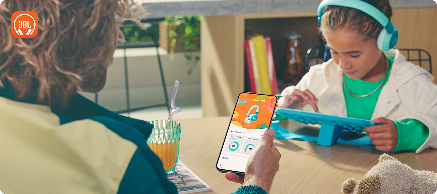 제품을 착용한 여자아이가 태블릿 PC를 만지고 있습니다. 여자 아이 앞에 앉은 성인이 스마트 폰으로 Headphones 앱 화면을 보고 있습니다.