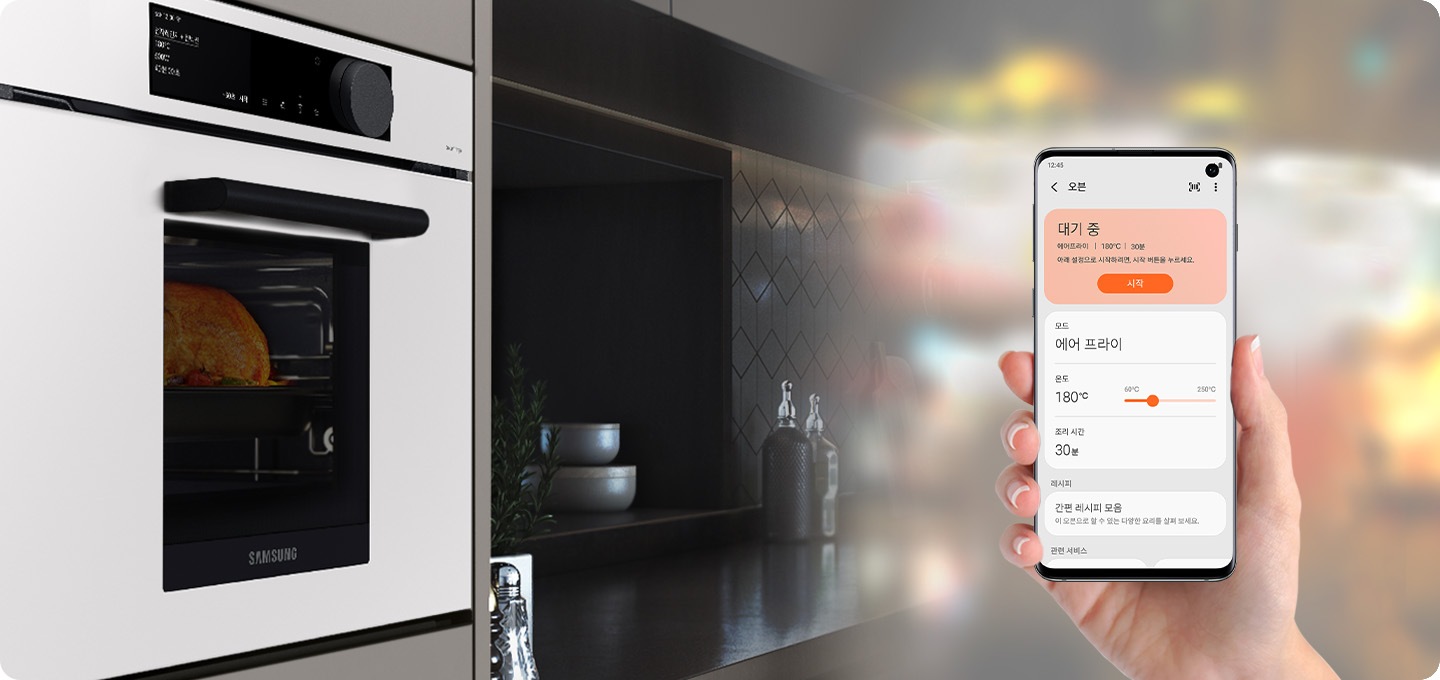 블랙 대리석 인테리어의 주방이 측면으로 기울여져 보여지고 있습니다. 우측엔 글램 화이트 색상의 오븐이 있고 오븐 안에는 통닭이 연출되어 있습니다. 가운데는 블랙 대리석 인테리어의 주방에 사기 그릇 2개가 쌓여 있습니다. 좌측엔 블러처리로 된 주방 인테리어 위에 손으로 스마트폰을 잡은 상태에서 스마트폰의 화면이 보여집니다. 해당 화면은 스마트 싱스의 오븐을 원격 조정 할 수 있는 UI화면이 보여집니다.