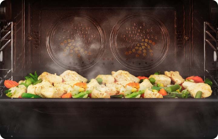 오븐이 작동되고 있는 연출된 이미지 입니다. 오븐 내에 듀얼 팬으로 가열 기능이 작동하고 있으며, 오븐 내부의 오븐 판 위에는 익혀지고 있는 닭과 각종 채소들이 보여집니다. 