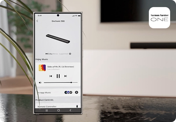 스마트폰에 Harman Kardon One 앱 화면이 보입니다. 스마트폰 뒤로는 TV와 사운드바가 흐릿하게 보입니다.