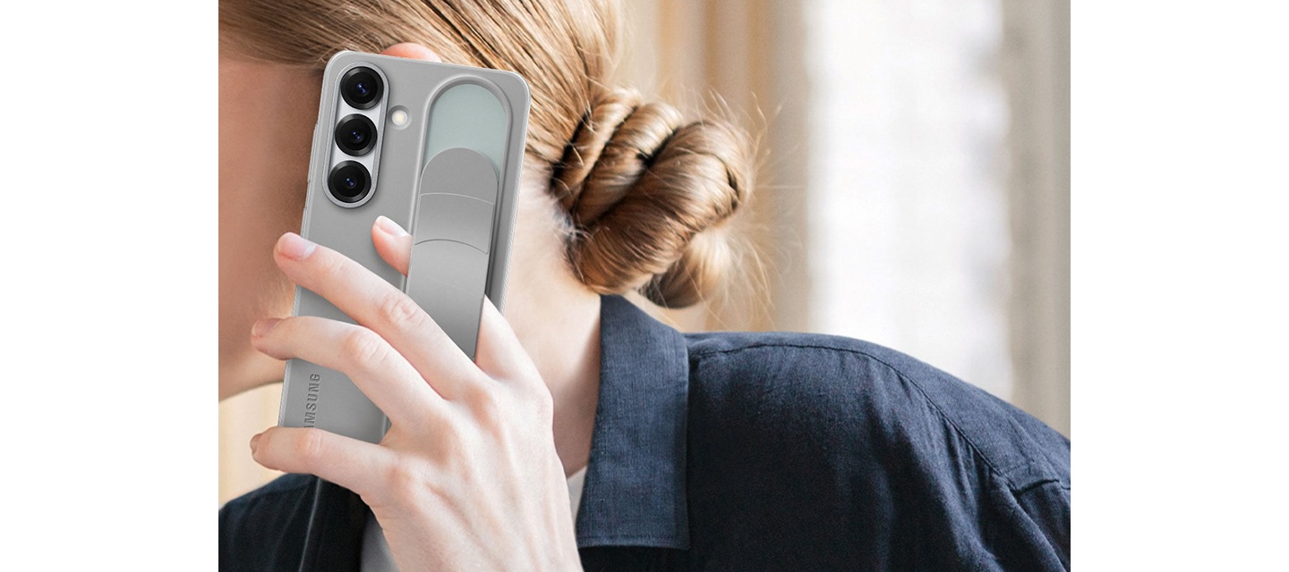 한 여성이 그레이 색상의 스탠딩 그립 케이스가 장착된 갤럭시 S25를 들고 있습니다. 한 손가락이 그립 아래에 끼워져 있습니다.