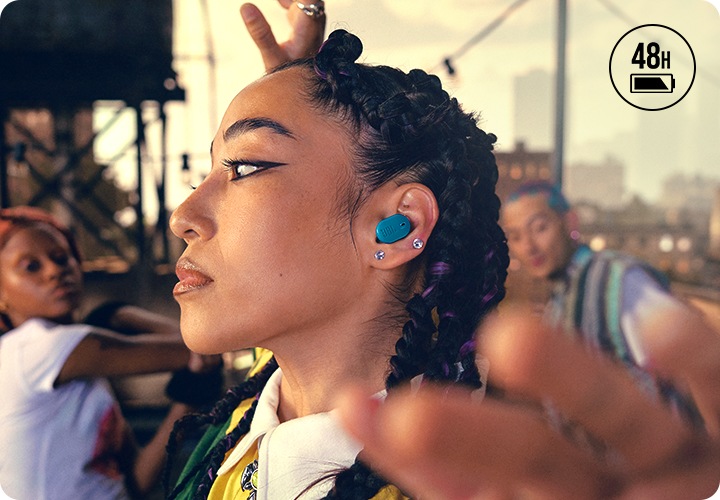 블루 색상의 이어버드를 착용한 여성이 보입니다. 뒤에는 춤을 추고 있는 사람이 보입니다.