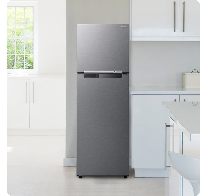 화이트톤의 주방 인테리어컷 입니다. 냉장고 좌측에 창문이 있고 밖으로는 초록색 나뭇잎이 보이며, 아래에 흰색 하부장이 있습니다. 중앙에 냉장고가 놓여있고 우측으로는 화이트톤의 상하부장이 있고 다양한 오브제가 놓여있습니다.