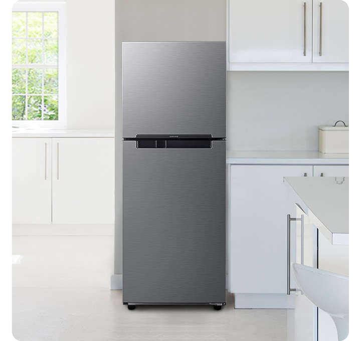 화이트톤의 주방 인테리어컷 입니다. 냉장고 좌측에 창문이 있고 밖으로는 초록색 나뭇잎이 보이며, 아래에 흰색 하부장이 있습니다. 중앙에 냉장고가 놓여있고 우측으로는 화이트톤의 상하부장이 있고 다양한 오브제가 놓여있습니다.