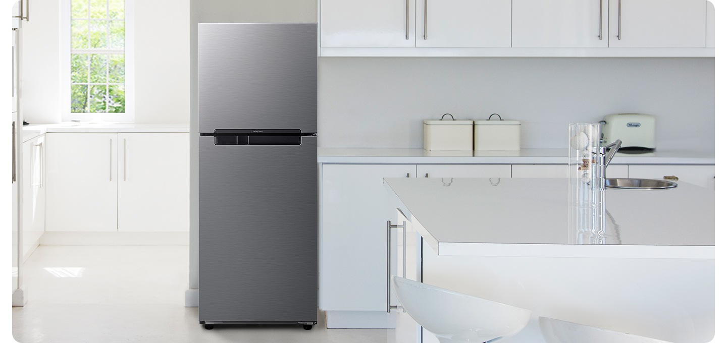 화이트톤의 주방 인테리어컷 입니다. 냉장고 좌측에 창문이 있고 밖으로는 초록색 나뭇잎이 보이며, 아래에 흰색 하부장이 있습니다. 중앙에 냉장고가 놓여있고 우측으로는 화이트톤의 상하부장이 있고 다양한 오브제가 놓여있습니다.