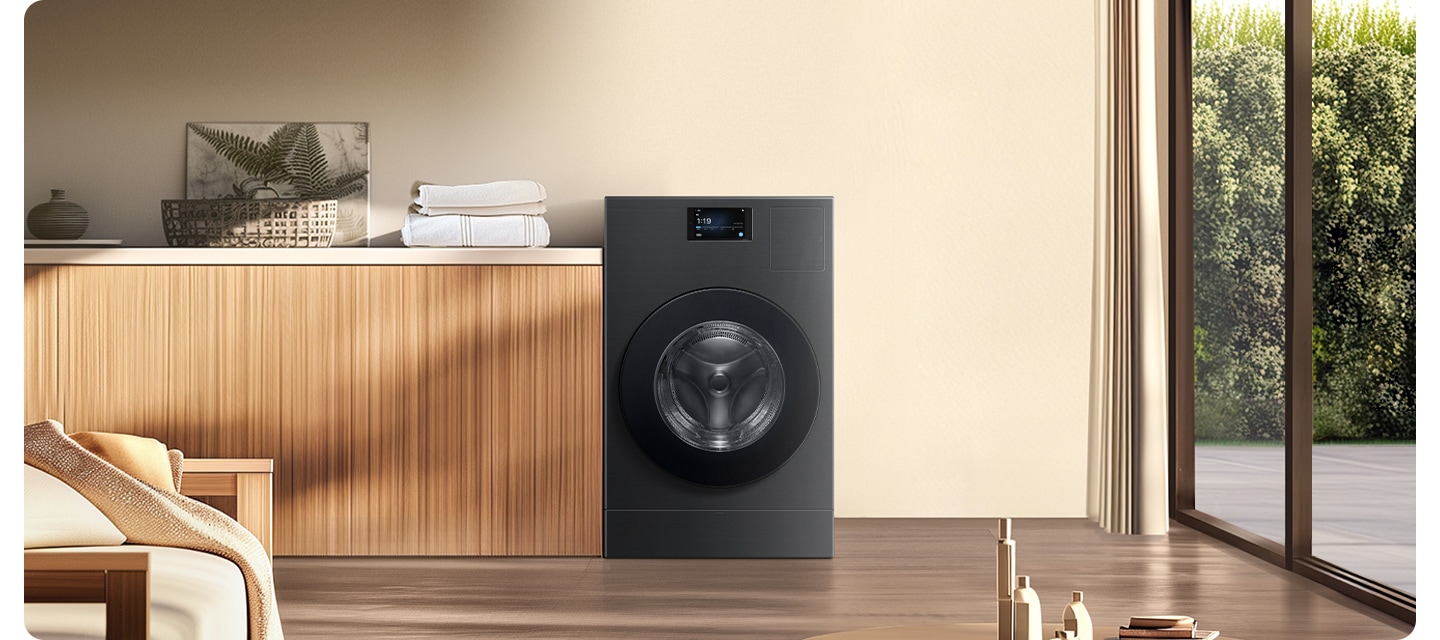 따뜻한 우드톤 거실에 세탁기 있습니다. 세탁기 왼쪽에는 아일랜드 탁자있고 위에는 그림, 바구니, 수건 등이 있습니다. 그 위에는 상단 수납공간도 있고 다양한 물건이 있습니다. 세탁기 오른쪽에는 큰 창이 있습니다, 창 밖에는 푸른나무가 보입니다.