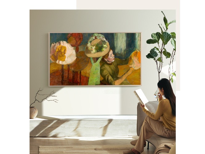 여성이 거실 소파에 앉아서 책을 보며 음료를 마시고 있습니다. 벽에는 TV가 설치되어 있으며, 화면에는 명화가 보입니다.