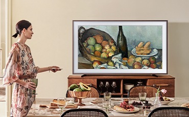 식탁 옆에 여성이 서서 TV를 보고 있습니다. TV 화면에는 명화가 보입니다. 식탁 위에는 과일과 유리컵이 놓여 있습니다..