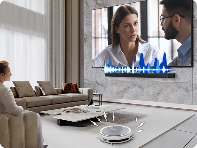 거실에서 여성이 TV를 보고 있습니다. TV 화면에는 드라마에 한 장면이 나오고 있습니다. TV 하단에는 사운드바가 설치되어 있고 TV와 사운드바 사이에는 음향효과가 있습니다. 거실 바닥에는 무선 로봇 청소기가 움직이고 있습니다.