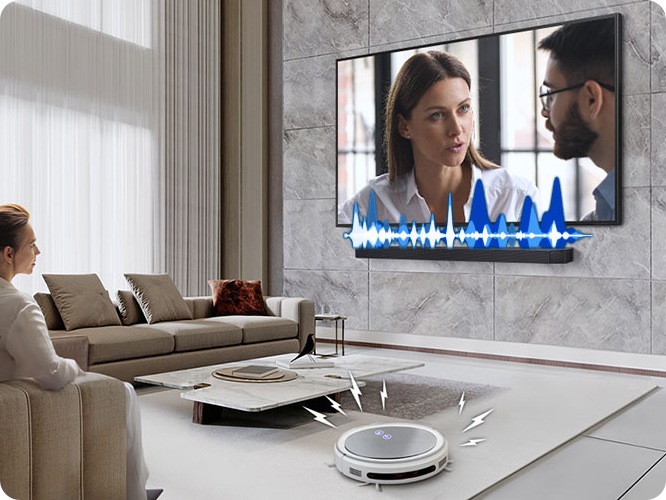 거실에서 여성이 TV를 보고 있습니다. TV 화면에는 드라마에 한 장면이 나오고 있습니다. TV 하단에는 사운드바가 설치되어 있고 TV와 사운드바 사이에는 음향효과가 있습니다. 거실 바닥에는 무선 로봇 청소기가 움직이고 있습니다.