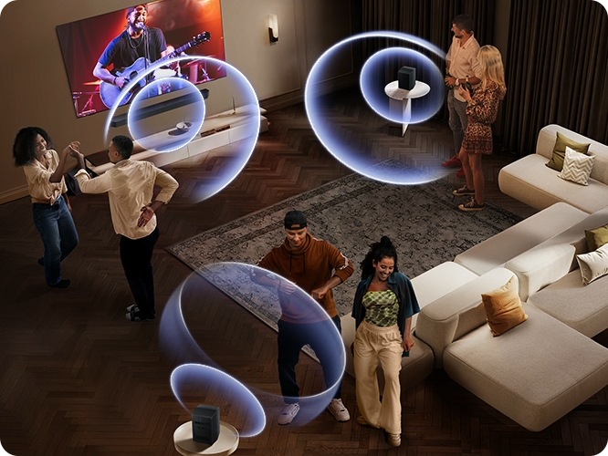 거실 벽에 TV와 사운드바가 설치되어 있습니다. TV 화면에는 공연장에서 사람들이 공연을 즐기고 있습니다. 사운드바와 소파 뒤에 연결된 리어 스피커에서 음향효과가 나오고 있습니다. 사람들이 거실 중앙에서 춤을 추고 있습니다.