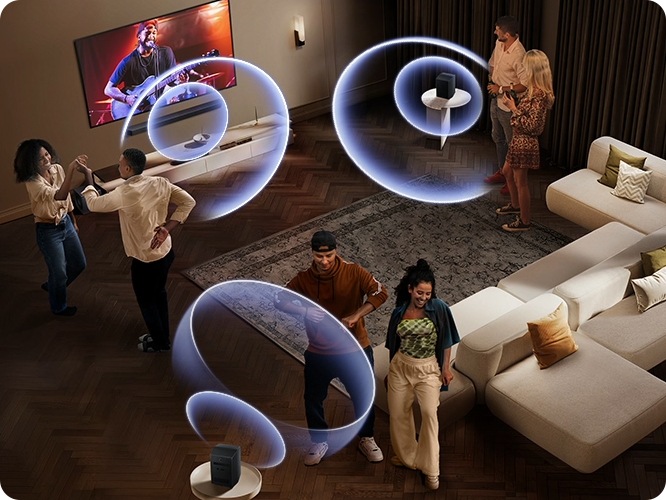 거실 벽에 TV와 사운드바가 설치되어 있습니다. TV 화면에는 공연장에서 사람들이 공연을 즐기고 있습니다. 사운드바와 소파 뒤에 연결된 리어 스피커에서 음향효과가 나오고 있습니다. 사람들이 거실 중앙에서 춤을 추고 있습니다.