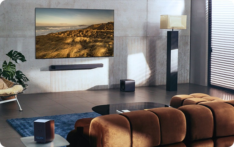 벽에 TV와 사운드바가 설치되어 있습니다. 사운드바 오른쪽 아래에는 서브 우퍼와 조명이 있으며, 소파 왼쪽 옆에는 무선 리어 스피커가 있습니다.