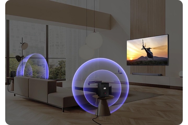 벽에 TV가 설치되어 있으며, 화면에는 헬리콥터가 보입니다. TV 앞에는 소파와 테이블이 있습니다. 소파 양옆에는 무선 리어스피커가 설치되어 있으며, 파란색 음향효과가 나오고 있습니다.