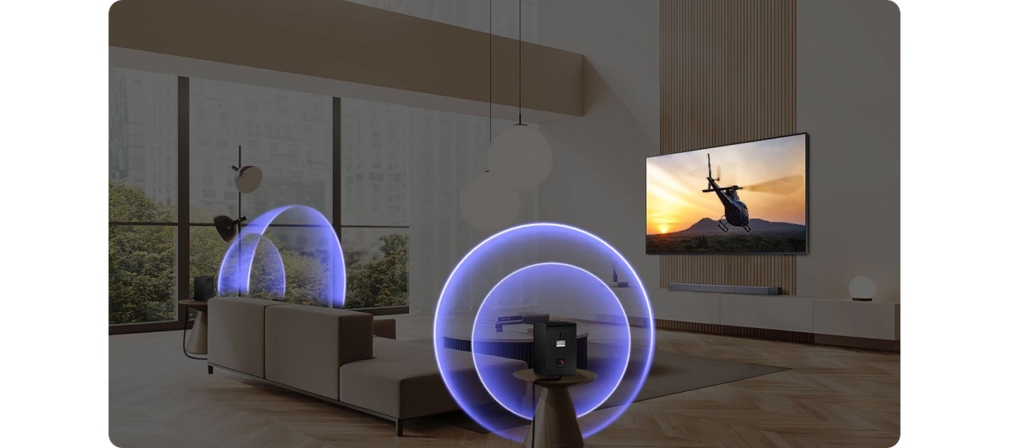 벽에 TV가 설치되어 있으며, 화면에는 헬리콥터가 보입니다. TV 앞에는 소파와 테이블이 있습니다. 소파 양옆에는 무선 리어스피커가 설치되어 있으며, 파란색 음향효과가 나오고 있습니다.