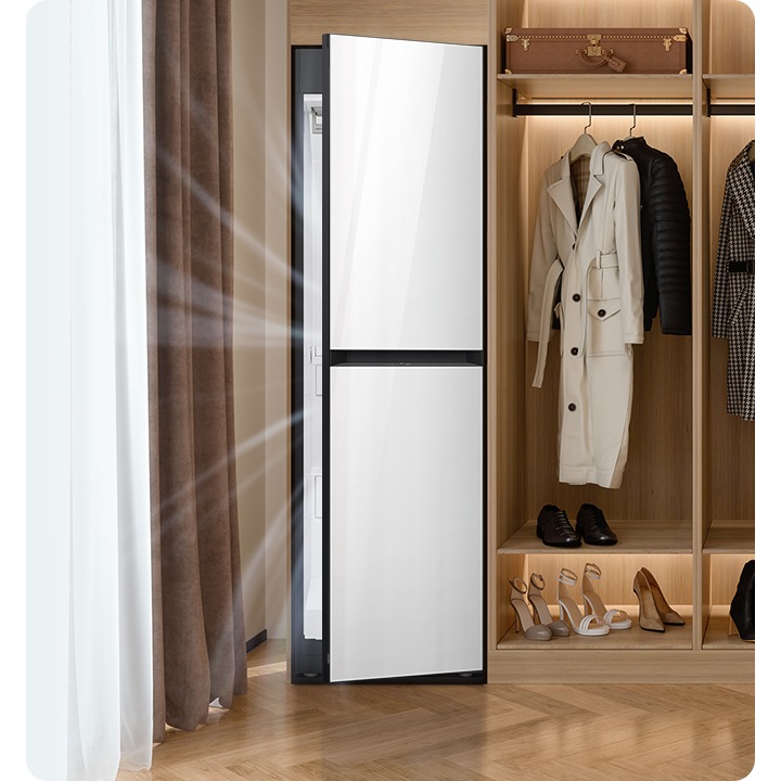 따뜻한 톤의 행거옷장이 좌측에 있고 옷걸이에는 옷이 두벌 걸려있습니다, 옷 위로는 캐리어 가방 있고, 아래에는 여러 개의 구두있습니다. 바로 옆 우측에는 에어드레서 드레스룸을 제습하고 있는 모습입니다.