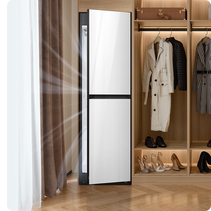 따뜻한 톤의 행거옷장이 좌측에 있고 옷걸이에는 옷이 두벌 걸려있습니다, 옷 위로는 캐리어 가방 있고, 아래에는 여러 개의 구두있습니다. 바로 옆 우측에는 에어드레서 드레스룸을 제습하고 있는 모습입니다.