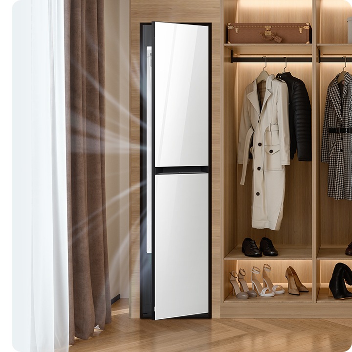 따뜻한 톤의 행거옷장이 좌측에 있고 옷걸이에는 옷이 두벌 걸려있습니다, 옷 위로는 캐리어 가방 있고, 아래에는 여러 개의 구두있습니다. 바로 옆 우측에는 에어드레서 드레스룸을 제습하고 있는 모습입니다.