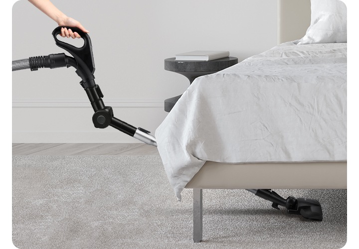 연한 회색 톤의 침실을 배경으로 이미지 오른쪽 부분에는 침대가 보이고 있고, 이미지 왼쪽 부분에는 엘보 연장관을 활용하여 침대 밑까지 청소하고 있는 모습이 보입니다.