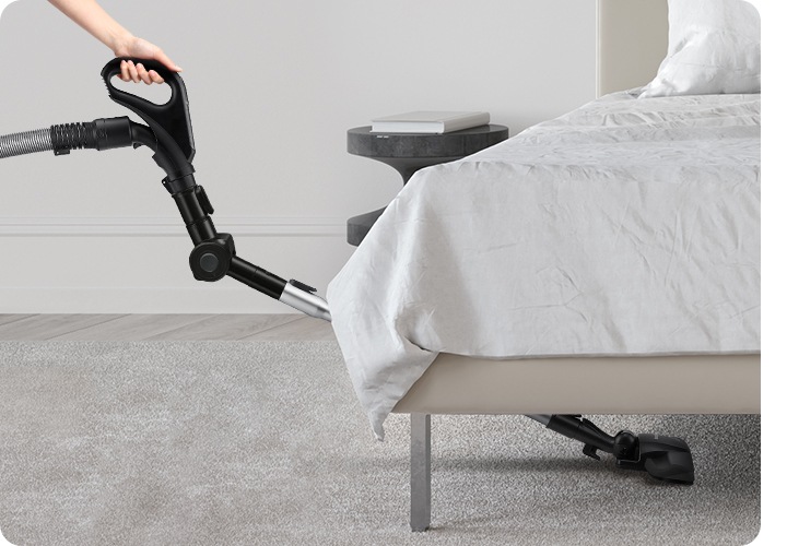 연한 회색 톤의 침실을 배경으로 이미지 오른쪽 부분에는 침대가 보이고 있고, 이미지 왼쪽 부분에는 엘보 연장관을 활용하여 침대 밑까지 청소하고 있는 모습이 보입니다.