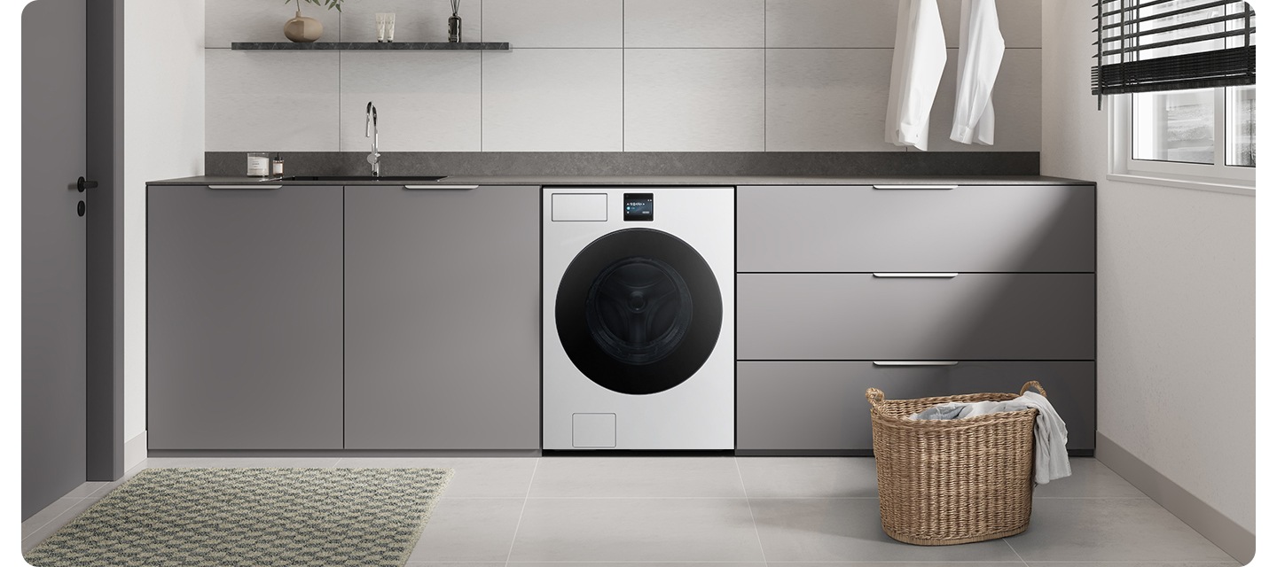 화이트&그레이톤 인테리어컷으로 그레이톤 하부장 중앙에 Bespoke AI 세탁기가 설치되어 있습니다. 상부장 부분에는 싱크대와 수전이 있고 앞쪽에는 라탄 바구니에 옷감이 들어있는 연출된 이미지입니다.