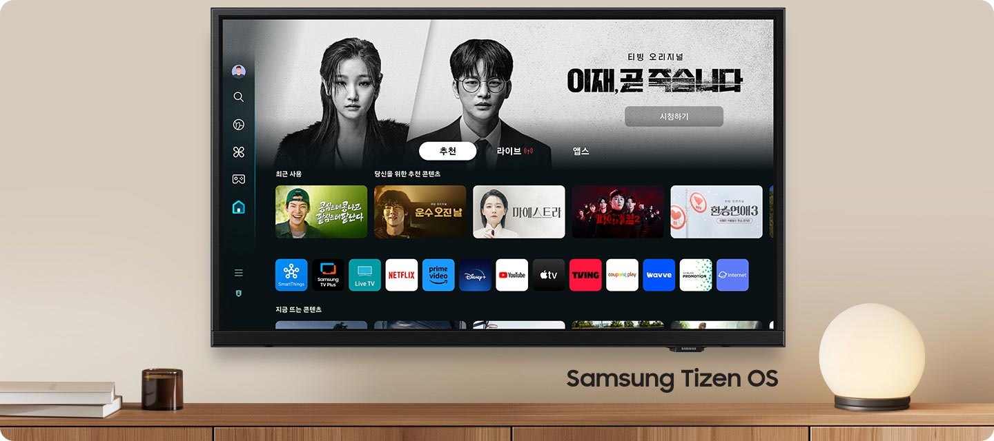 벽에 TV가 설치되어 있으며, 화면 속에는 이재 곧 죽습니다 드라마 메인 화면이 보이고 있으며, 그 하단에는 여러 예능과 드라만 메인 화면이 보입니다. TV 오른쪽 하단에는 Samsung Tizen OS 문구가 기재되어 있습니다.