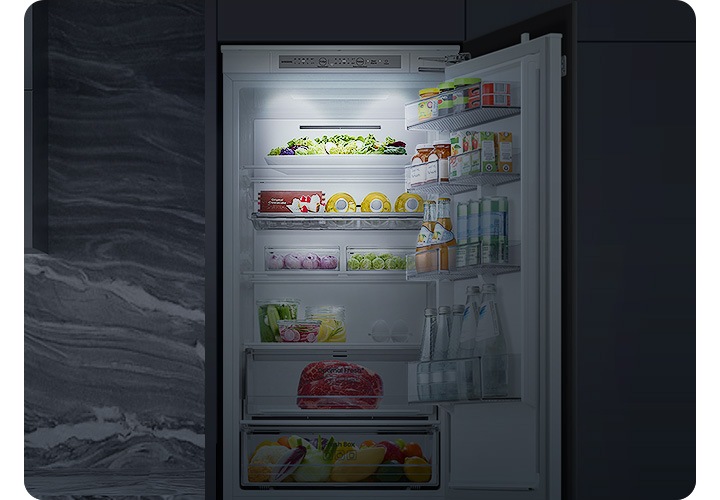 어두운 실내에 빌트인 냉장고가 설치되어 있고 도어가 오픈되어 있습니다. 도어 내부에서 은은한 조명이 냉장고 내부를 밝혀주는 연출된 이미지 입니다.