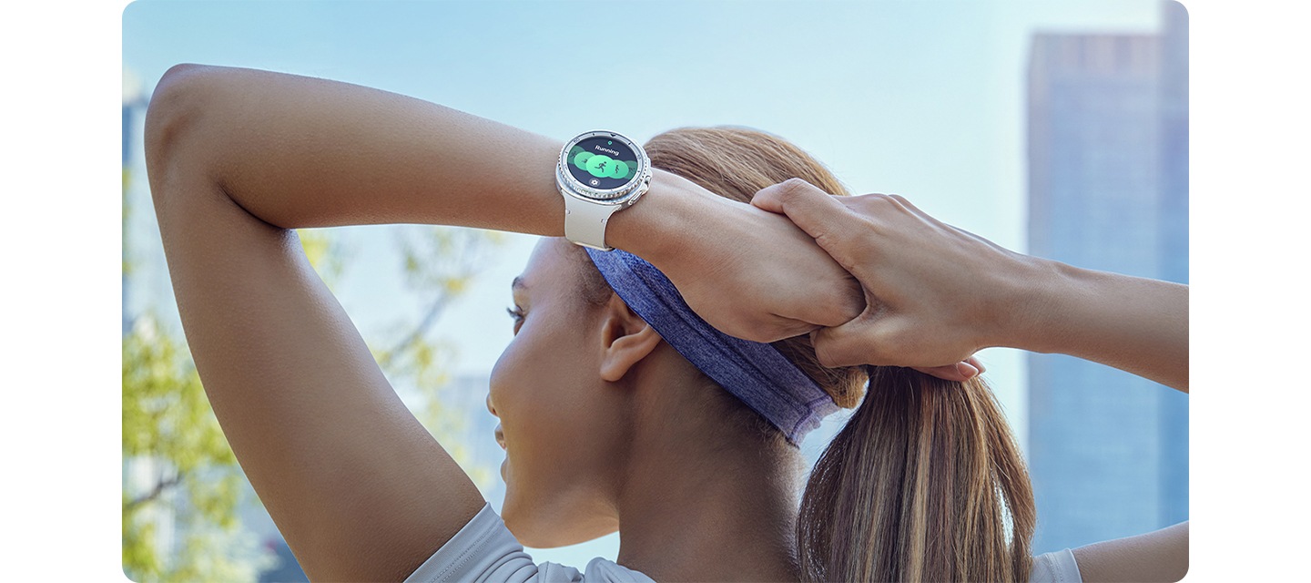 도시를 배경으로 여성이 머리 뒤로 팔을 구부려 스트레칭을 합니다. 손목 위 갤럭시 워치8 클래식에는 화이트 색상의 갤럭시 워치8 스포츠 밴드가 장착되어 있습니다.