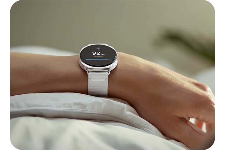 화이트 색상의 갤럭시 워치8 패브릭 밴드가 장착된 갤럭시 워치8을 착용한 사람이 이불 위에 팔을 올려놓고 있습니다.