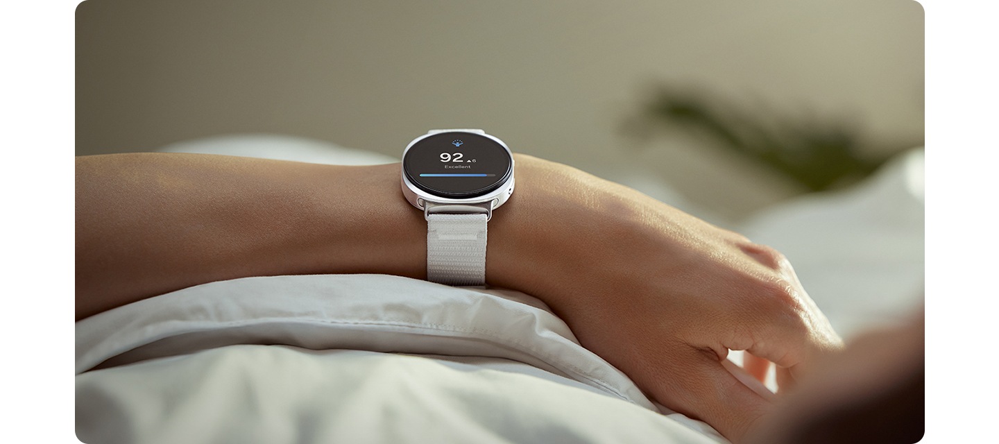 화이트 색상의 갤럭시 워치8 패브릭 밴드가 장착된 갤럭시 워치8을 착용한 사람이 이불 위에 팔을 올려놓고 있습니다.