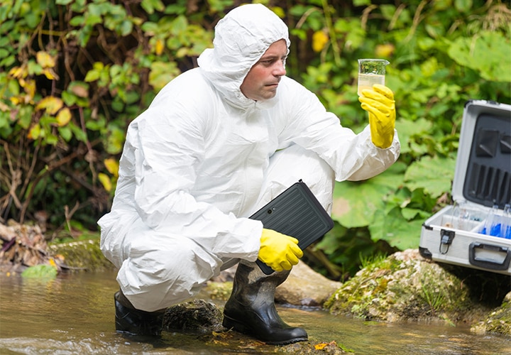 방진복과 보호 장비를 착용한 남성이 호숫가에서 수질 샘플을 채취하고 있습니다. 왼손에는 채취한 물이 담긴 비커, 오른손에는 갤럭시 탭 액티브5 프로를 들고 쪼그려앉아 있습니다. 기기가 물, 먼지 및 오염물에 강한 저항성을 지니고 있음을 명확하게 표현하고 있습니다.
