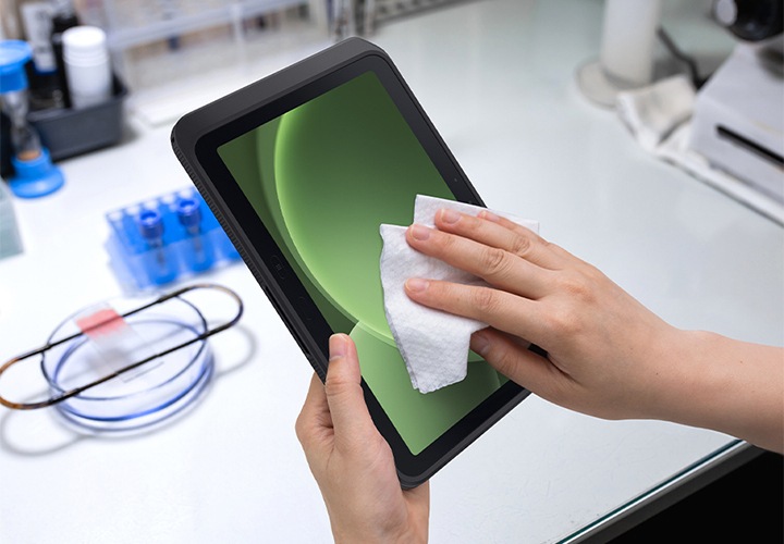 실험실 환경에서 갤럭시 탭 액티브5 프로를 세척하는 모습입니다. 왼손은 갤럭시 탭 액티브5 프로를 잡고 있으며 오른손에는 천을 들고 화면을 닦고 있습니다. 디스플레이에는 녹색 음영의 그래픽이 띄워져 있습니다.