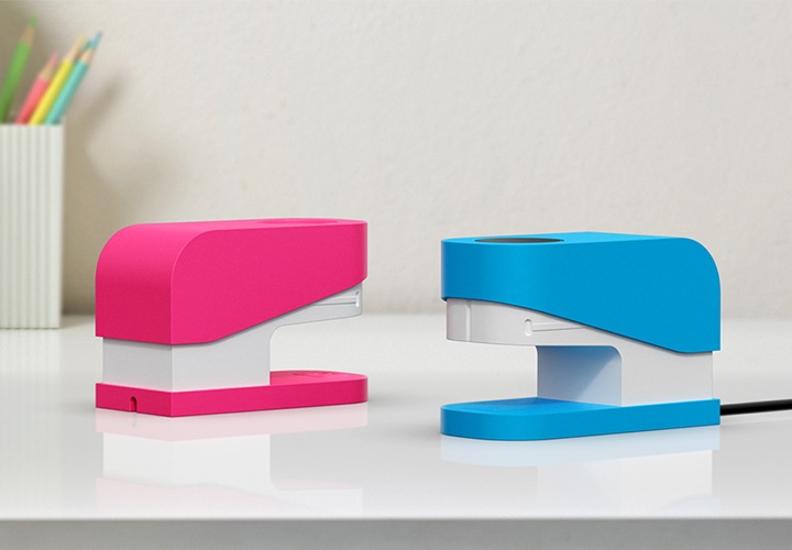 책상 위에 연필꽂이가 보이며 그 앞 왼쪽에 분홍색 스테이플러 갤럭시 워치 충전독 거치대가, 오른쪽에 충전독을 장착한 파란색 스테이플러 갤럭시 워치 충전독 거치대가 보이고 있습니다.