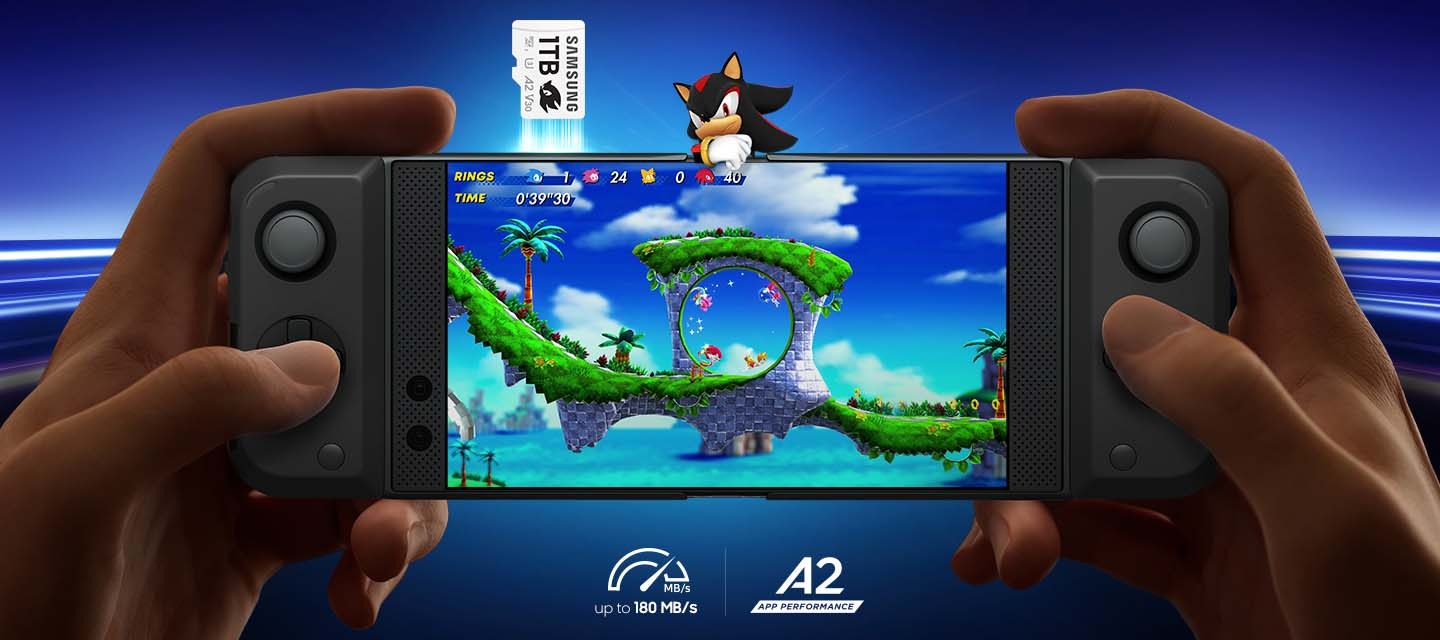 파란색 배경에 가상공간과 같은 곳에서 손으로 게임기를 잡고 게임기 안에 소닉 게임을 하고 있는 모습이 연출되고 있습니다. 게임기 위에는 섀도우가 게임기에 중간의 상단 부분에 팔을 걸치고 잇습니다. 섀도우 왼쪽으로 섀도우 마이크로SD 메모리카드 소닉 PRO Plus 1TB가 게임기 밖으로 튀어오르고 있습니다. 이미지 내 하단에는 up to 180 MB/s 아이콘과 A2 app performance 아이콘이 순서대로 보여집니다. 