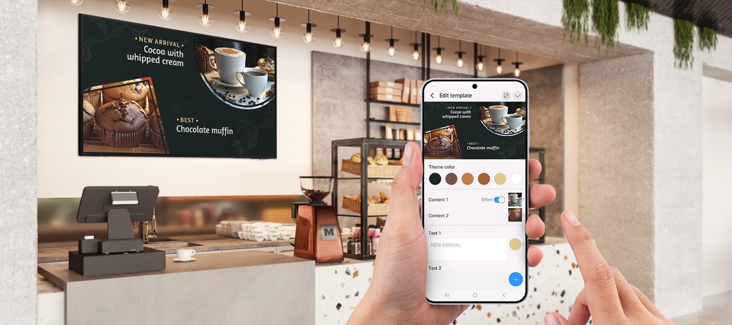 벽면에 비즈니스TV가 설치된 카페의 카운터를 바라보고 있으며, 그 앞으로 스마트폰을 들고 있는 손이 보입니다. 스마트폰 화면에는 설치된 비즈니스TV의 화면을 App으로 제어할 수 있는 화면이 보입니다.