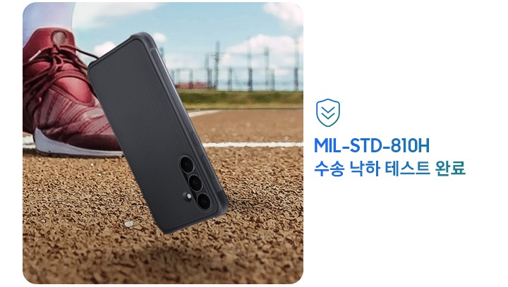 러기드 케이스가 장착된 갤럭시 S25 FE가 야외 트랙 바닥에 떨어지기 직전의 모습으로 2.5cm 정도 공중에 떠 있어, 낙하 사고가 발생했음을 알려줍니다. 떨어지는 스마트폰 뒤로 러닝화를 신은 사람의 발이 있습니다. 이미지 우측에는 '미국 국방부 군사 표준 규격 MIL-STD-810H에 따른 테스트'라는 표시가 있습니다.