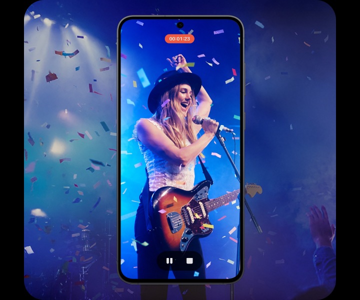 [갤럭시 S25 FÉ 화면에 라이브 무대 공연 녹화 영상이 표시됩니다. 검은색 모자를 쓴 공연자가 일렉트릭 기타를 연주하며 마이크에 대고 노래를 부릅니다. 파란색 무대 조명이 배경을 비추고 색종이 조각이 떨어집니다. 화면 상단에는 ‘00:01:23’ 타이머가 표시되어 있고 하단에는 재생 컨트롤이 있습니다.]