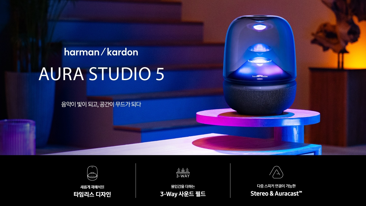 계단 앞 테이블 위에 스피커가 놓여 있으며, 스피커에서는 보라색 불빛이 나오고 있습니다. 스피커 뒤로는 화분들이 보입니다. 스피커 왼쪽으로는 하만카돈 로고와 AURA STUDIO 5 마크가 보입니다. 그 아래는 ‘음악이 빛이 되고, 공간이 무드가 되다’ 문구가 있습니다. 이미지 하단에는 ‘새롭게 재해석된 타임리스 디자인‘, ‘몰입감을 더하는 3-Way 사운드 필드‘, ‘다중 스피커 연결이 가능한 Stereo & Auracast™’ 문구와 아아콘이 있습니다.